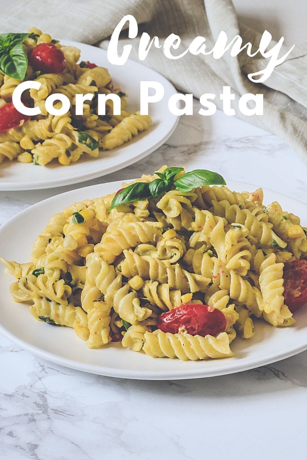Creamy corn pasta on a plate with garnish of basil leaves, another plate on back with napkin and text on top of the image