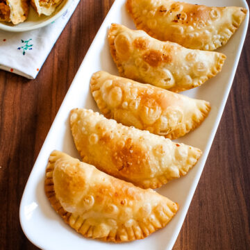 5 mawa gujiya in a plate with one cut into half in a small plate in the back with napkin underneath