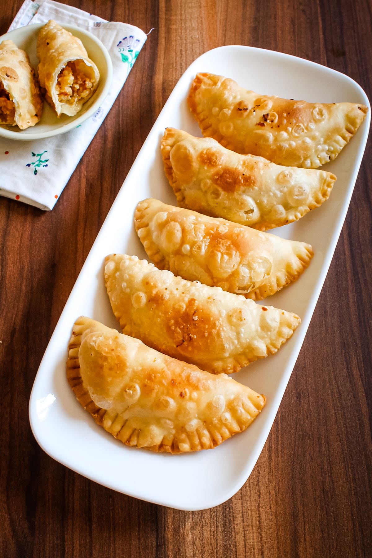 5 mawa gujiya in a plate with one cut into half in a small plate in the back with napkin underneath