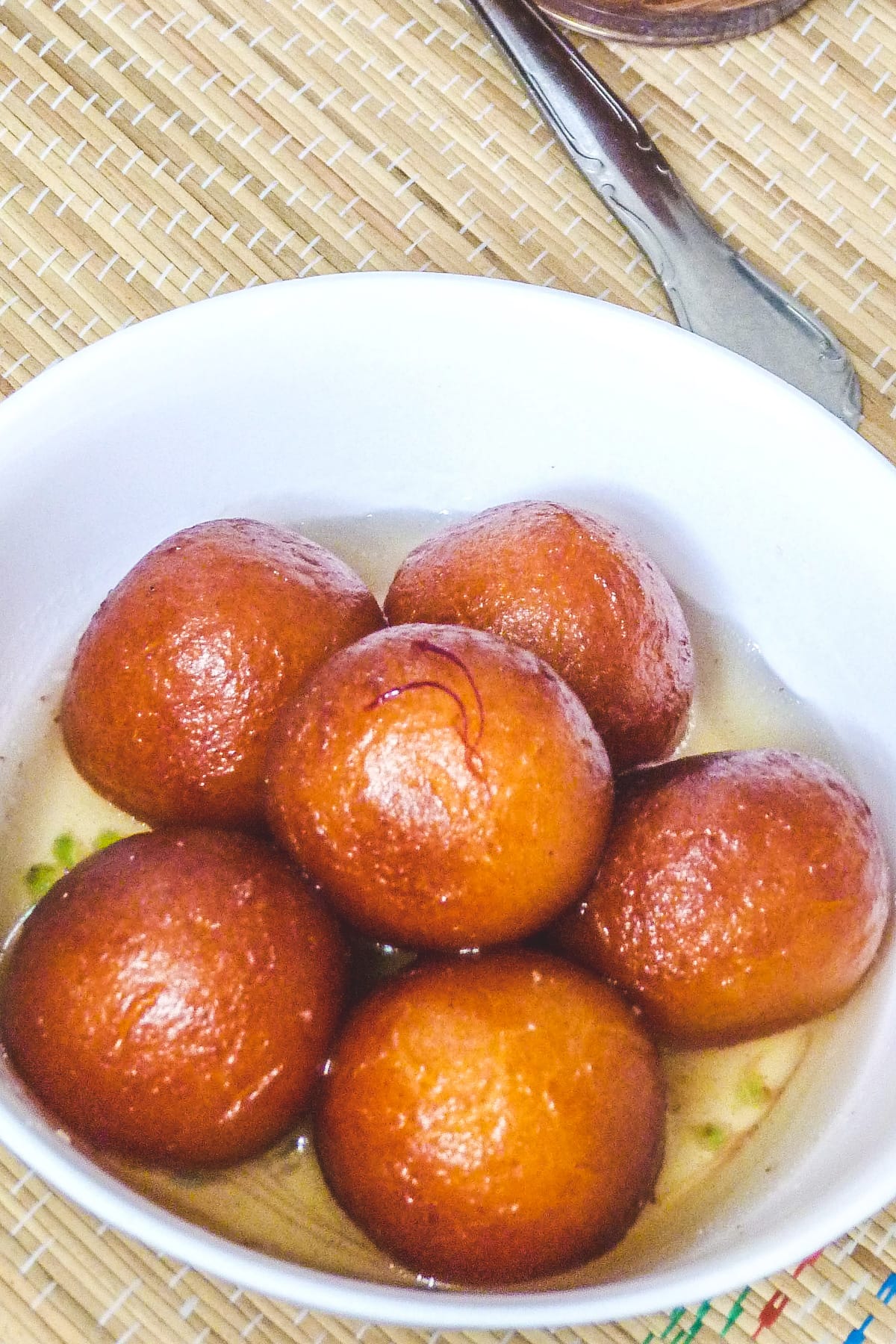 6 gulab jamun with sugar syrup in a bowl with spoon on side.