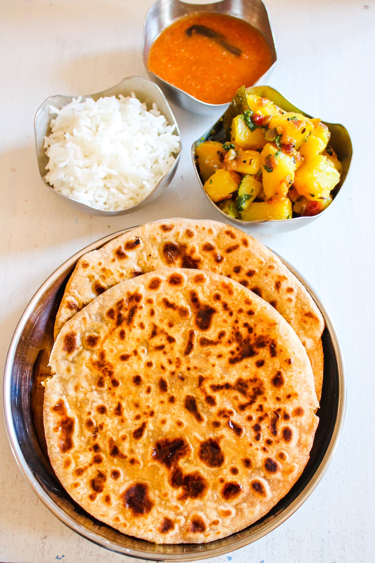 Two puran poli in a steel plate with rice, bhaji and amti in steel bowl on side.