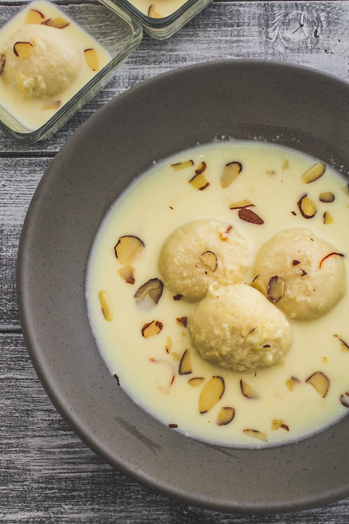 3 pieces of rasmalai in a gray bowl with garnish of almonds with one rasmalai in a small bowl in the back