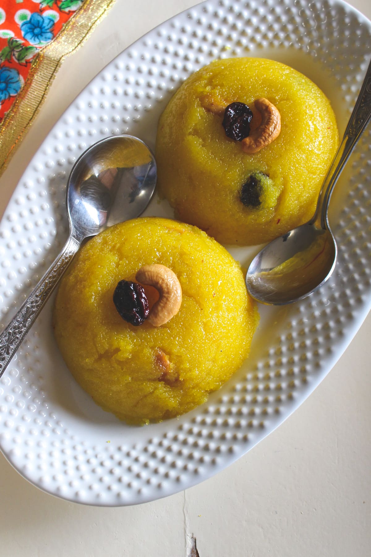 two scoops of rava kesari with garnish of cashew and raisin with 2 spoons in an oval plate