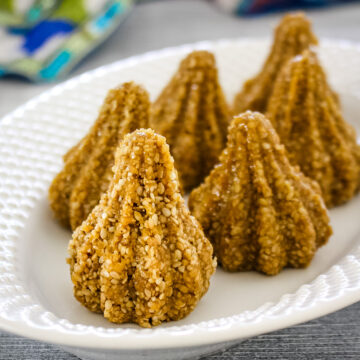 Close up of sesame seed modak in a white oval plate with colorful napkin in the back