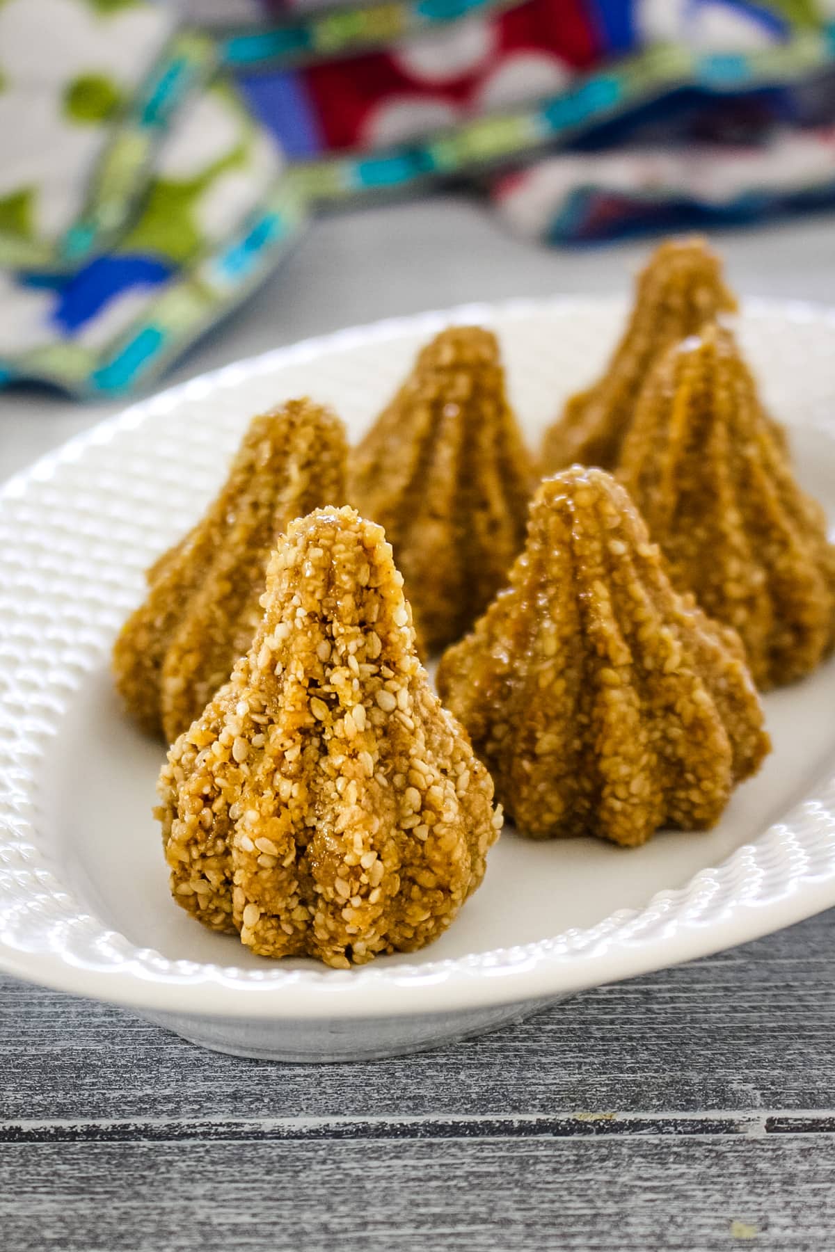 Close up of sesame seed modak in a white oval plate with colorful napkin in the back