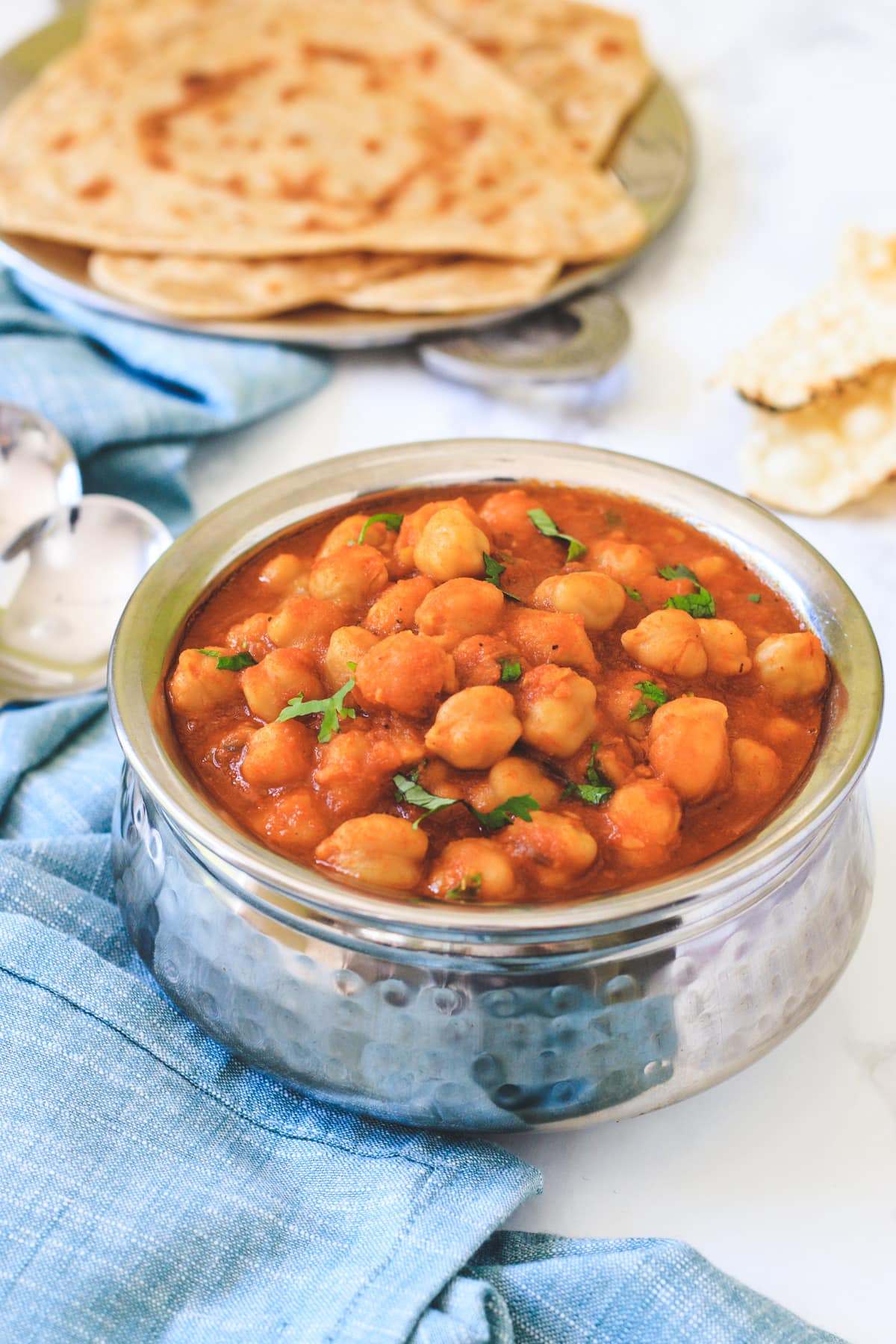 Jain chole in a steel handi with paratha served in the back, a napkin and spoons on the side.