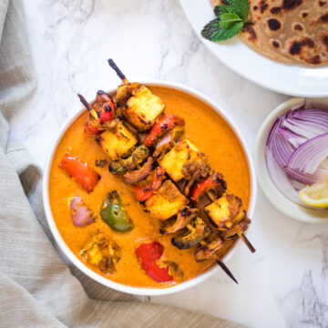 paneer tikka masala gravy with 2 skewers on the bowl, served with paratha, onions, chutney, lemon wedge