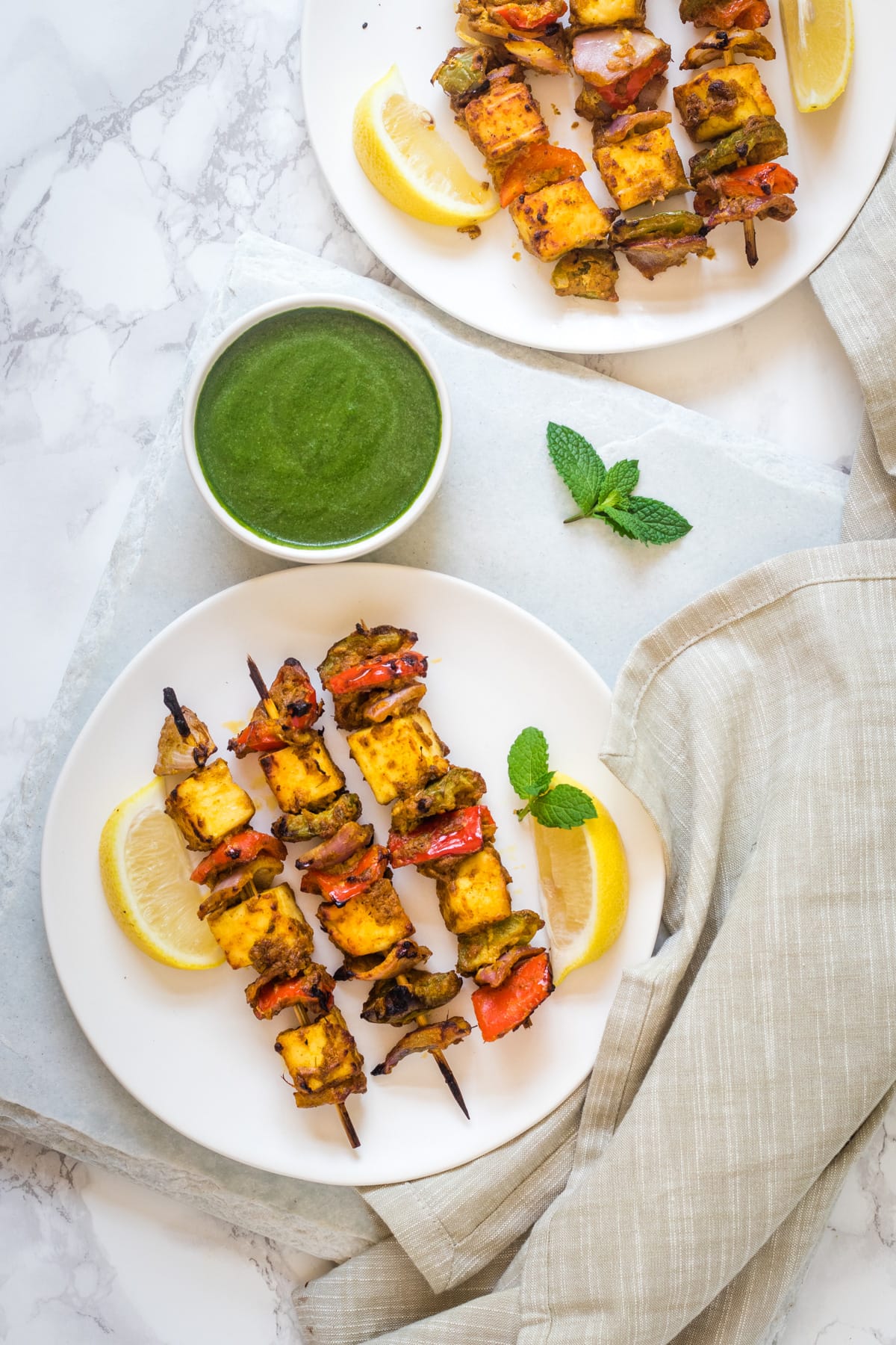 2 plates of paneer tikka served with lime wedges and green chutney with napkin on side.