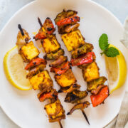 3 skewers of paneer tikka on a plate with lemon wedges and mint leaves in the plate, mint chutney in a bowl on side.