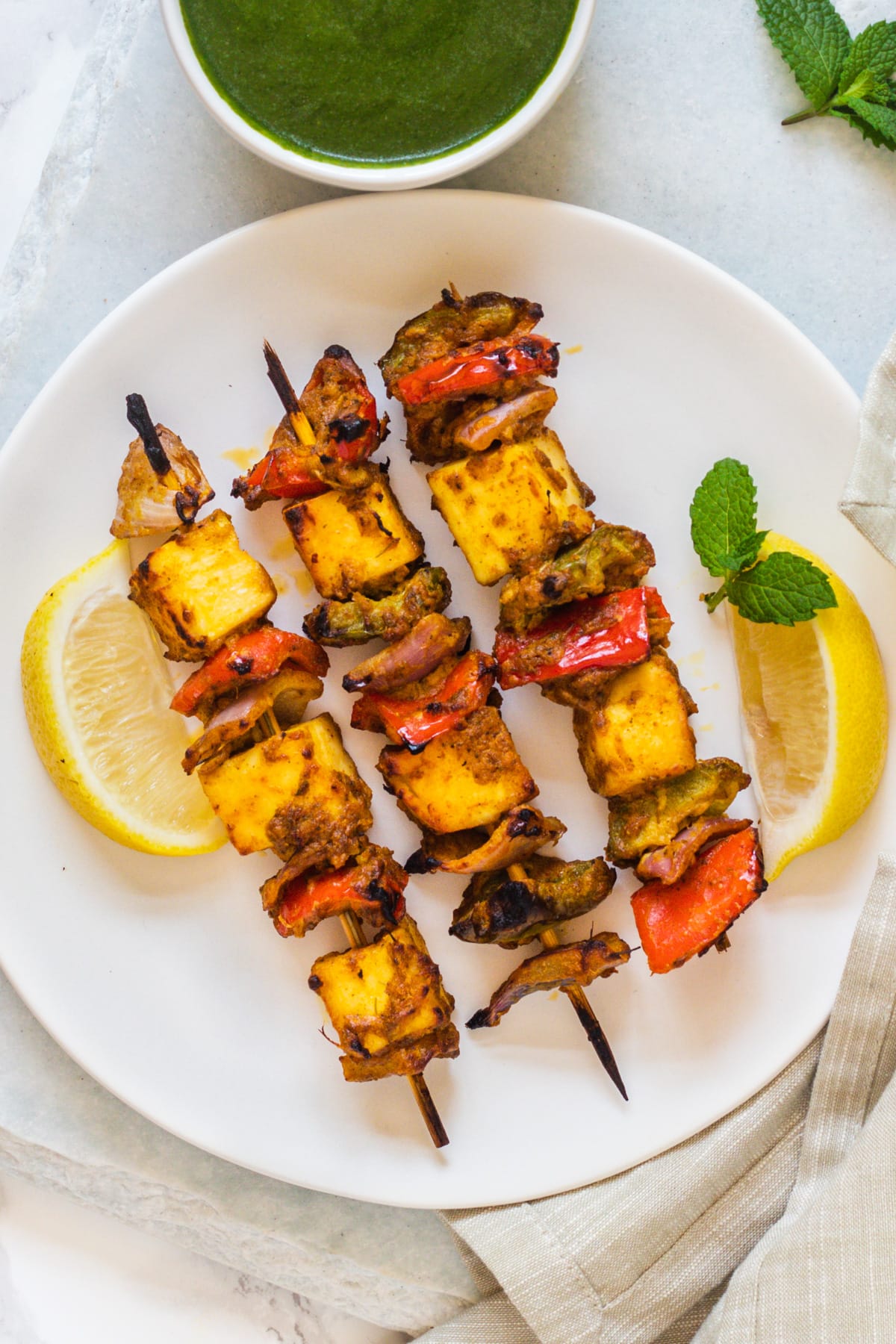 3 skewers of paneer tikka on a plate with lemon wedges and mint leaves in the plate, mint chutney in a bowl on side.