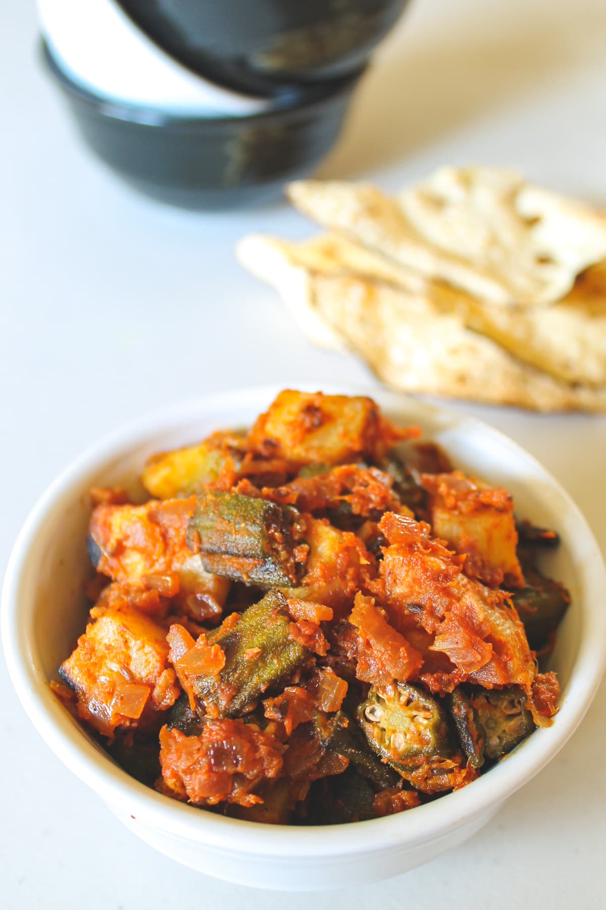 aloo bhindi served in a bowl with papad and stacked bowls in the back.