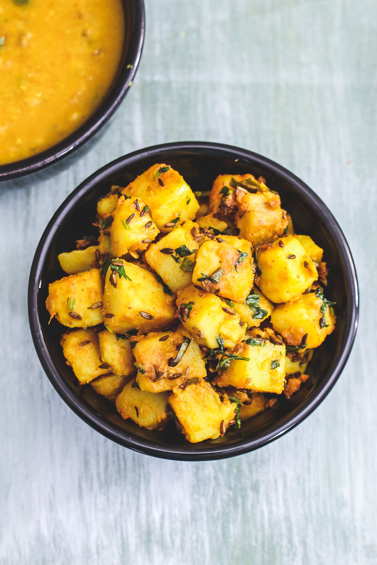 Top view of jeera aloo in a black bowl with dal served on side.