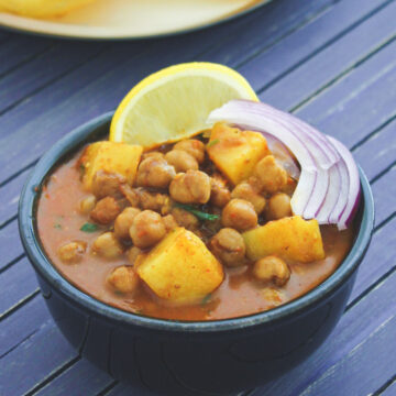 Close up of aloo chana in a black bowl with sliced onions and lemon wedge.