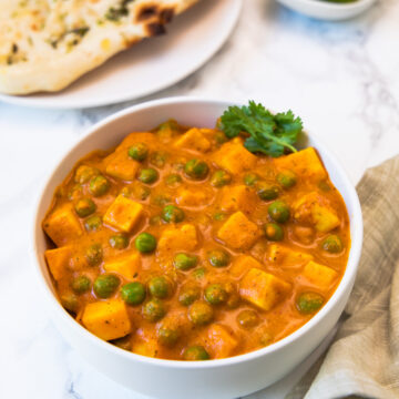 Matar paneer in a white bowl with side of naan, onions and lime wedges.