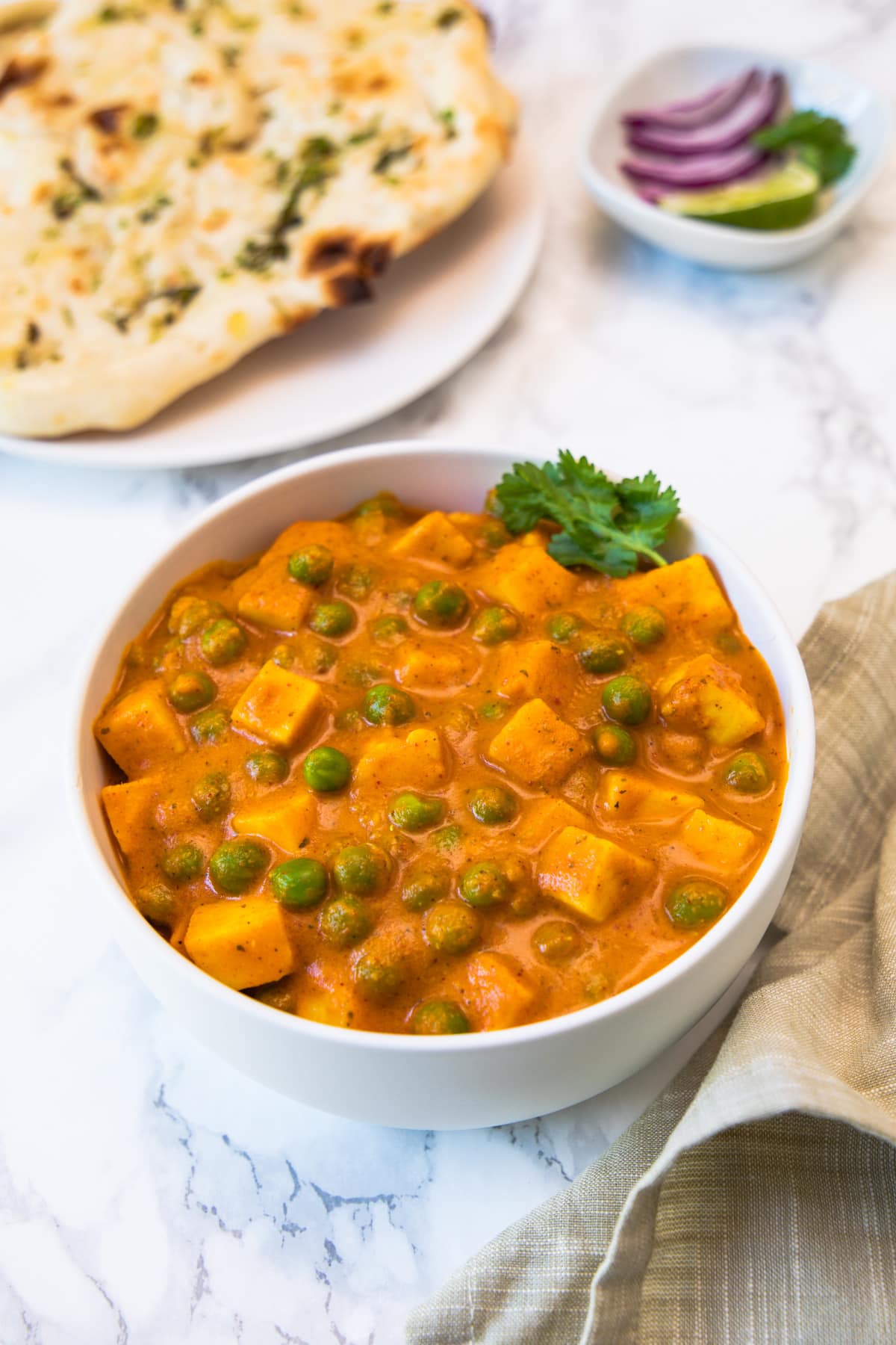 Matar paneer in a white bowl with side of naan, onions and lime wedges.