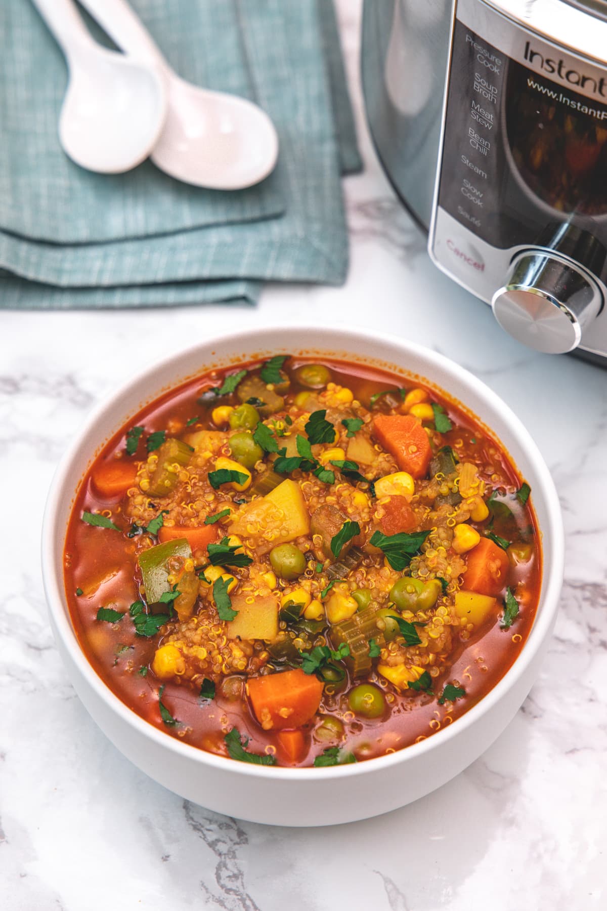 Quinoa vegetable soup served in a bowl with two spoons & napkin in the back.