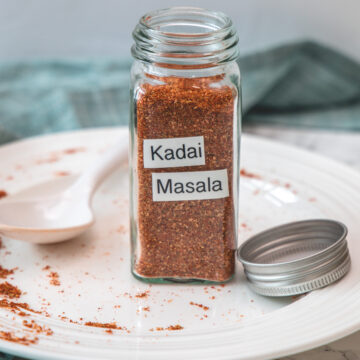 Kadai masala powder in a glass spice jar in a plate with spoon and lid with napkin on side.