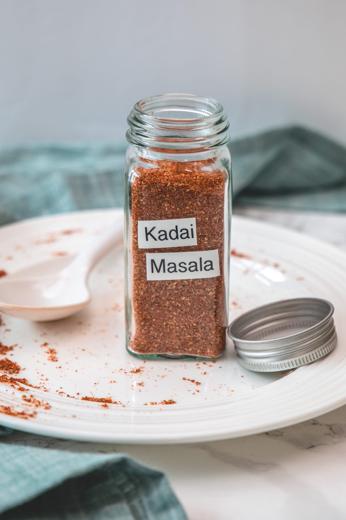 Kadai masala powder in a glass spice jar in a plate with spoon and lid with napkin on side.