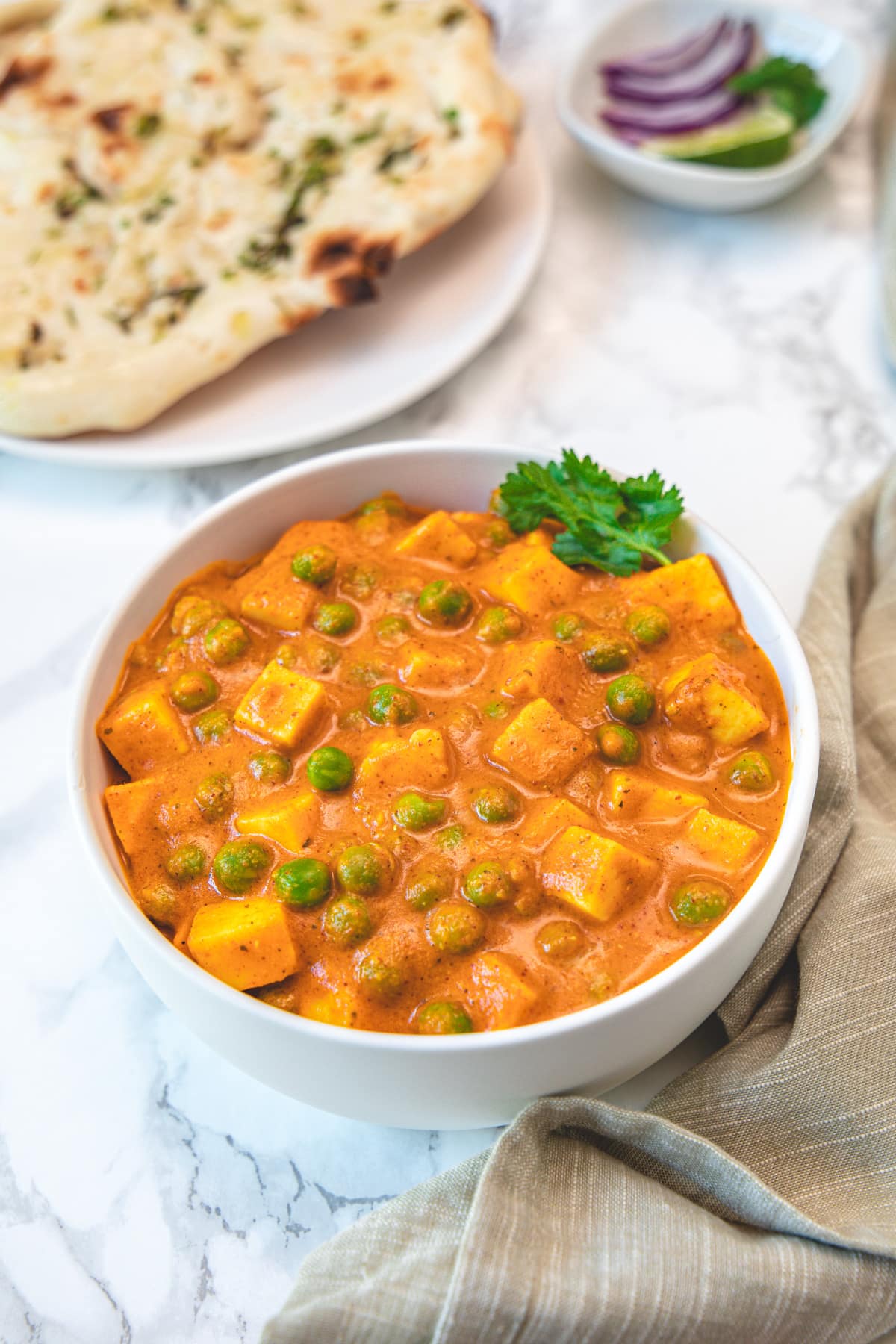 A bowl of matar paneer garnished with cilantro, naan and onions in the back.