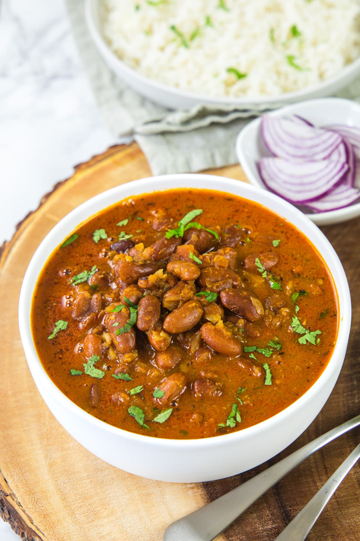 Rajma gravy served in a bowl garnished with cilantro, rice and onion in the back.