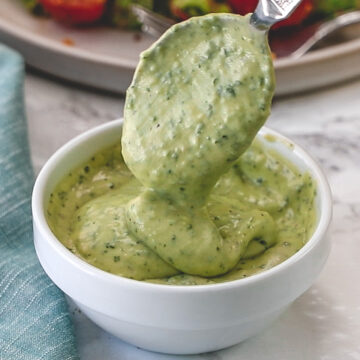 Drizzling a spoonful of dressing back into the bowl to show creamy texture with salad plate in back.