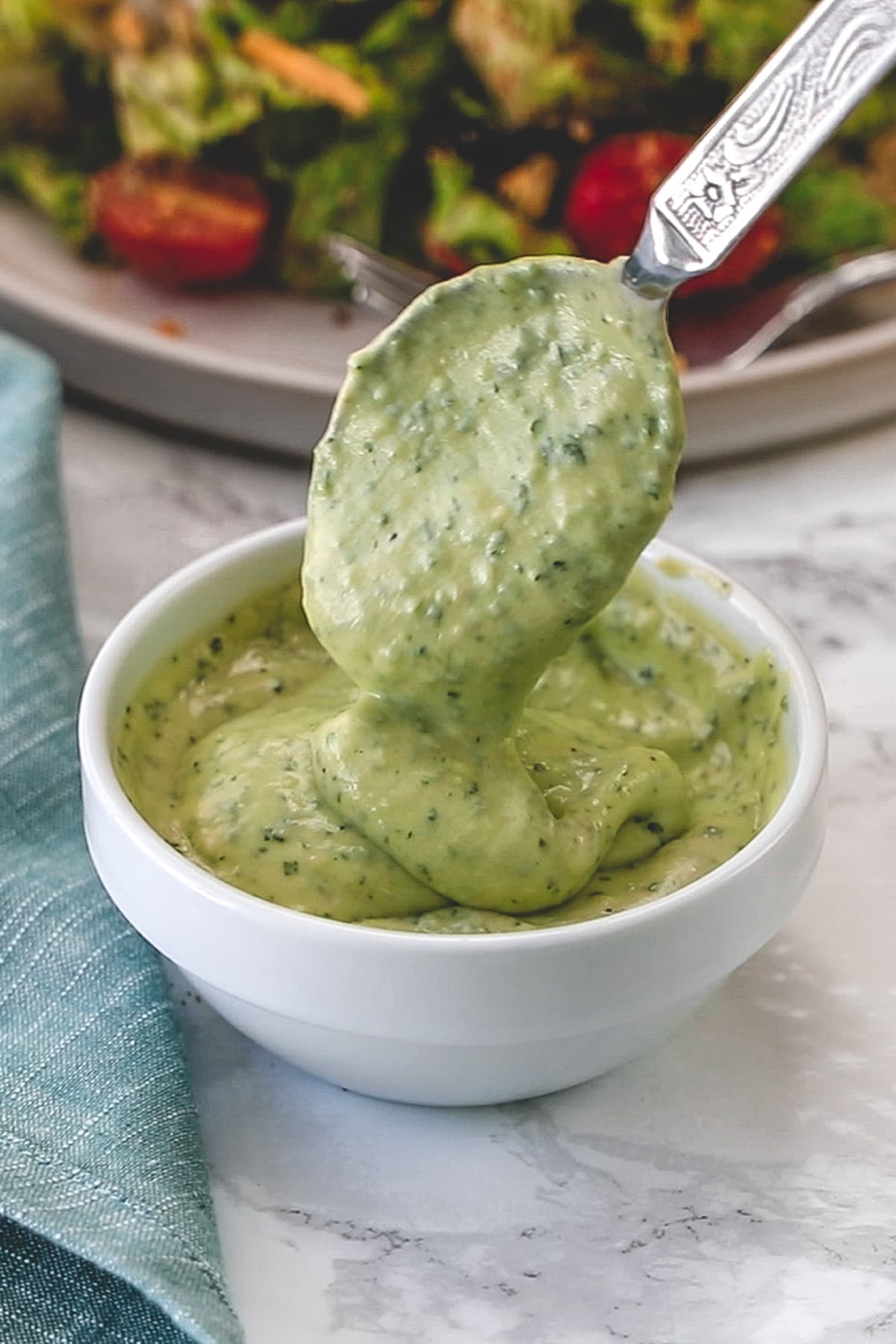 Drizzling a spoonful of dressing back into the bowl to show creamy texture with salad plate in back.