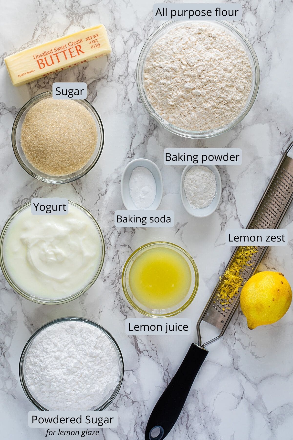 Ingredients used in eggless lemon cake in individual bowls on a marble surface with labels.