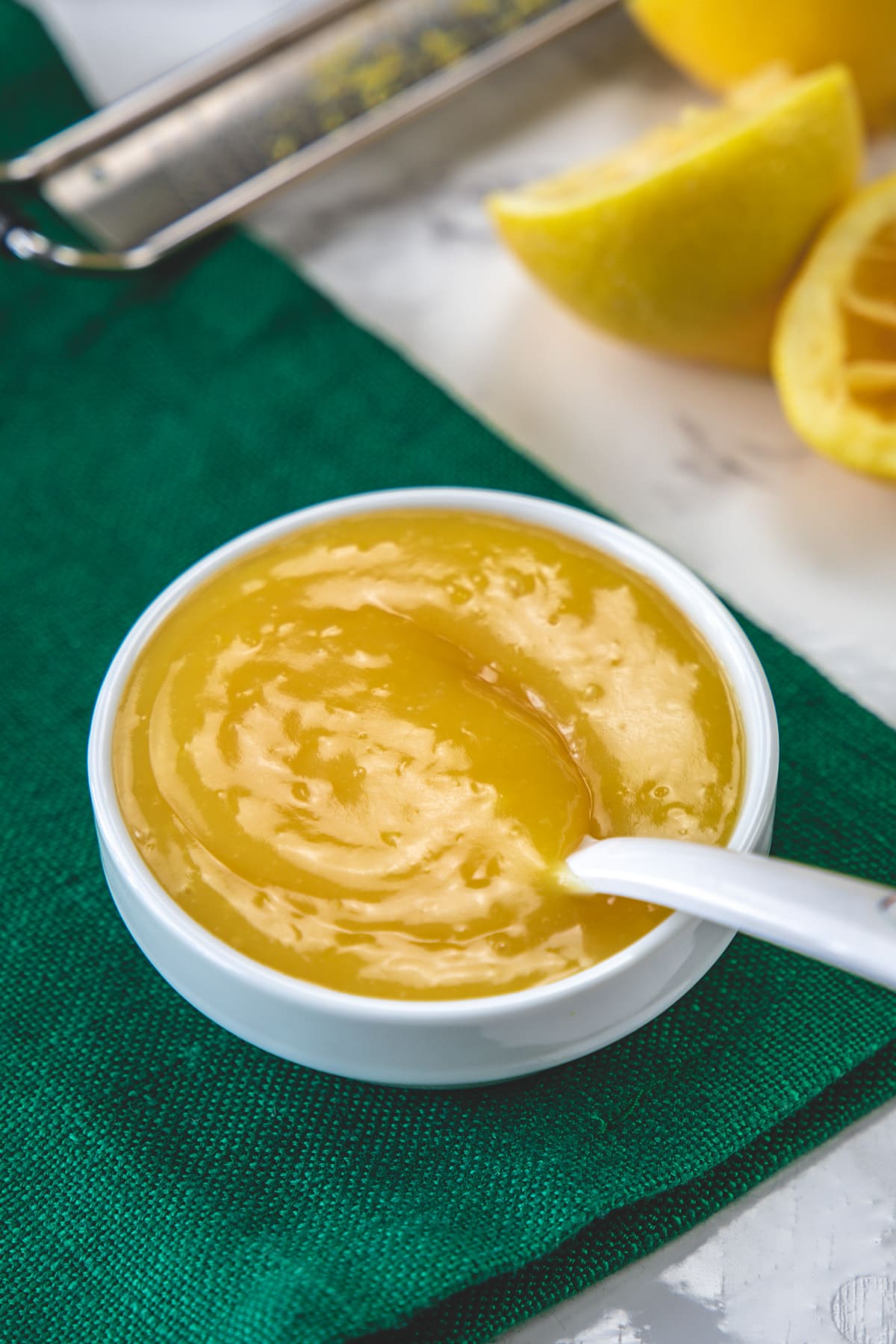 A bowl of eggless lemon curd on a green napkin with lemon halves and zester in the back.