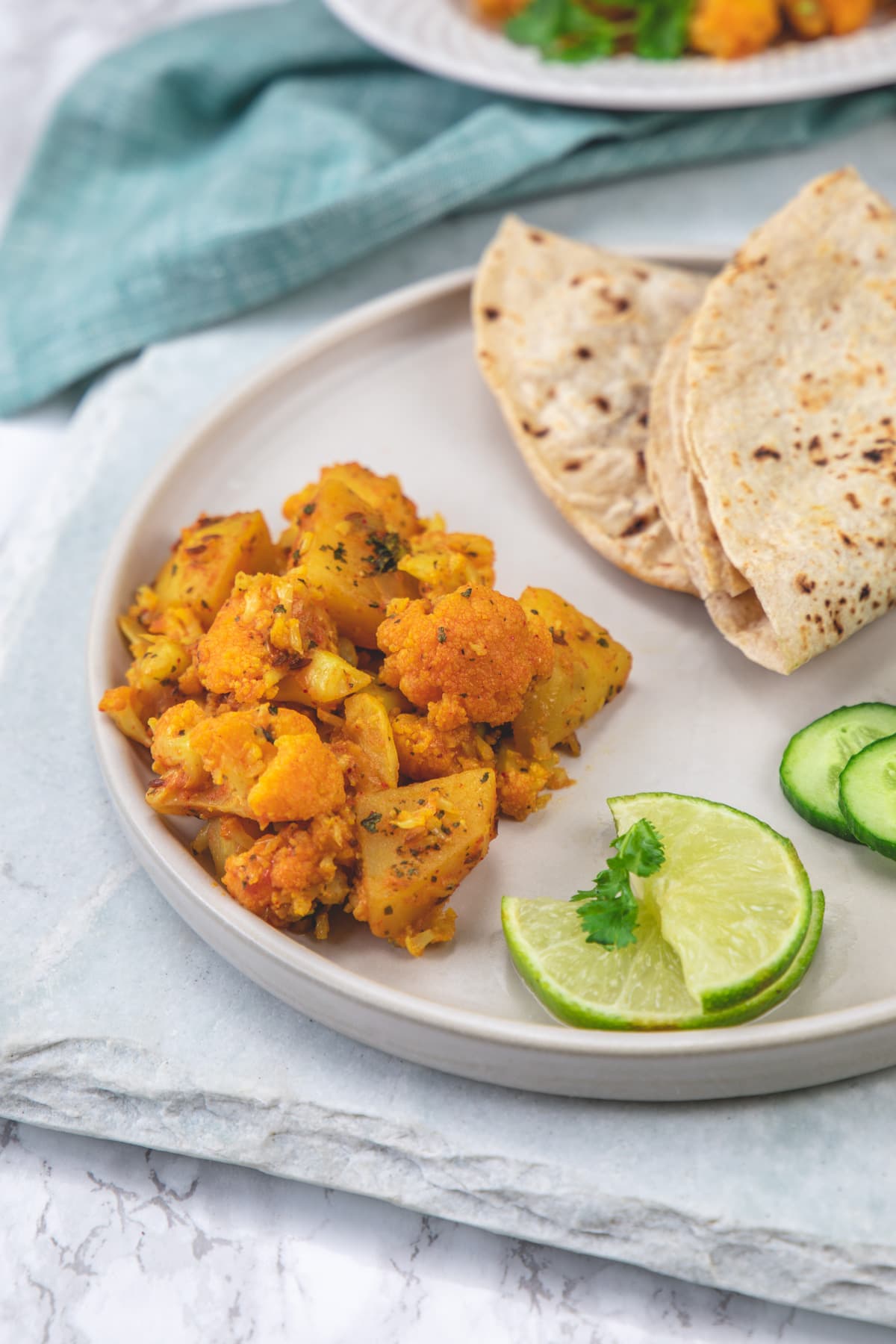 close up of aloo gobi served in a plate with roti, cucumber and lime.