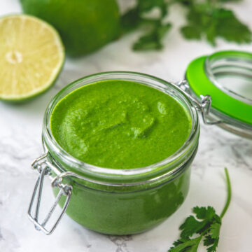 A glass jar of cilantro chutney with lime and cilantro in the back.