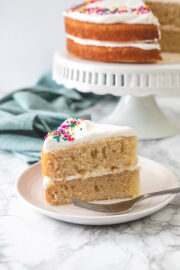 Eggless vanilla cake served in a plate with fork and remaining cake on cake stand in back.