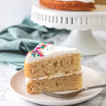 Eggless vanilla cake served in a plate with fork and remaining cake on cake stand in back.