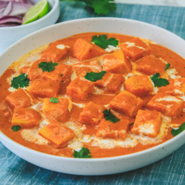 Paneer makhani garnished with cilantro, napkin under the bowl, onion, lime wedge and cilantro in the back.