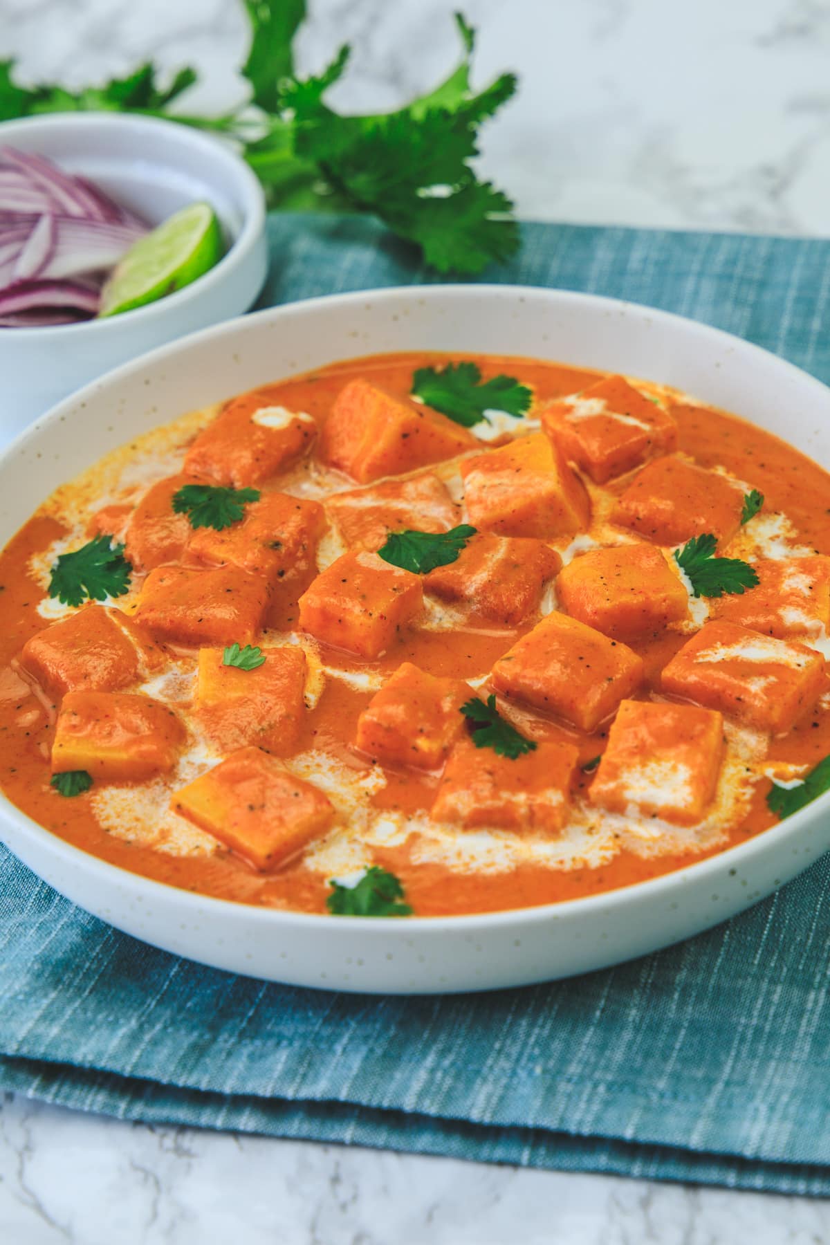 Paneer makhani garnished with cilantro, napkin under the bowl, onion, lime wedge and cilantro in the back.