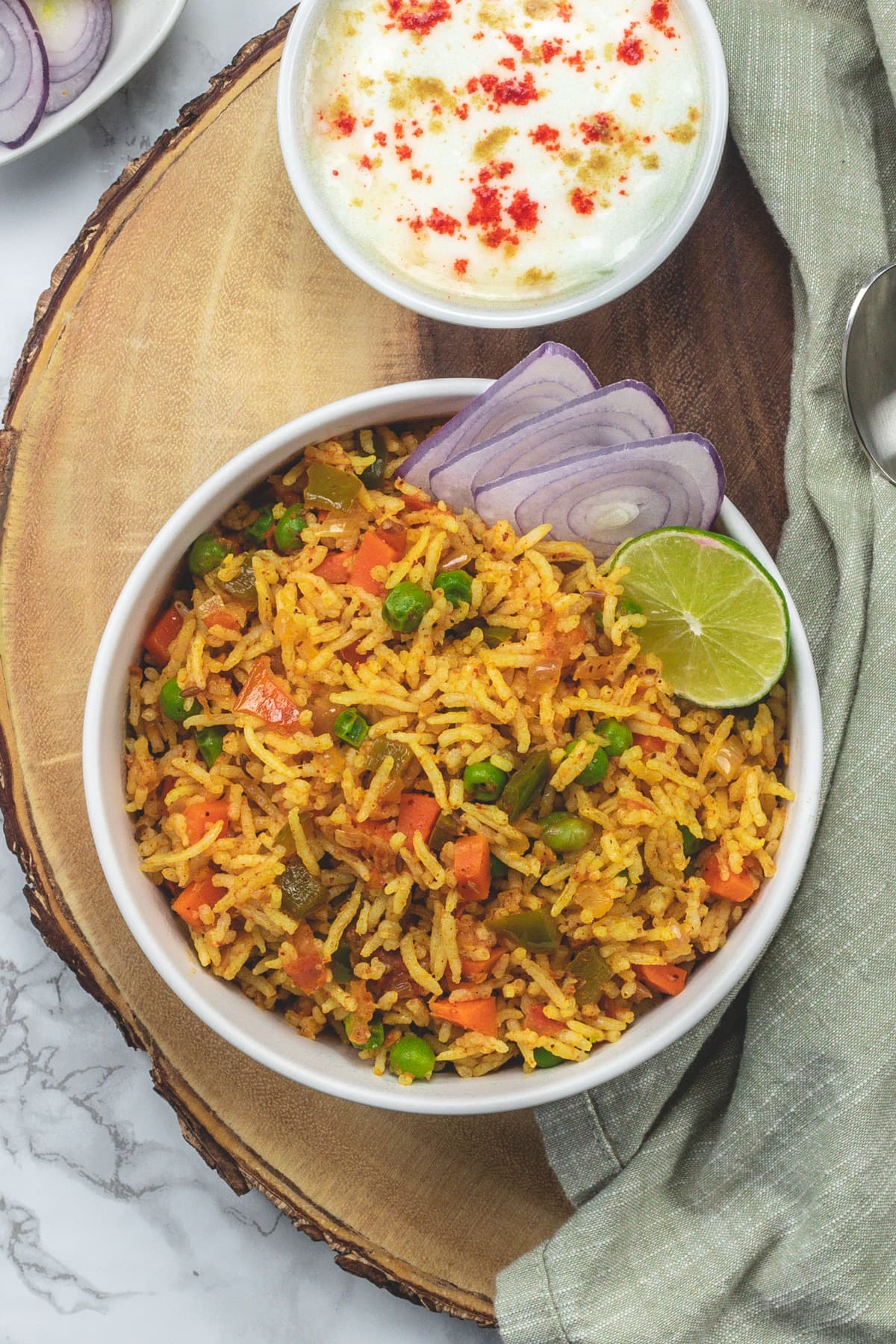 A bowl of tawa pulao garnished with onion slices, lime wedge and yogurt served on side.