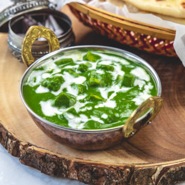 Palak paneer served in kadai with a garnish of cream and side of naan, onions.