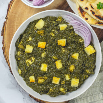 Saag paneer with sliced onions and naan on a wooden board.