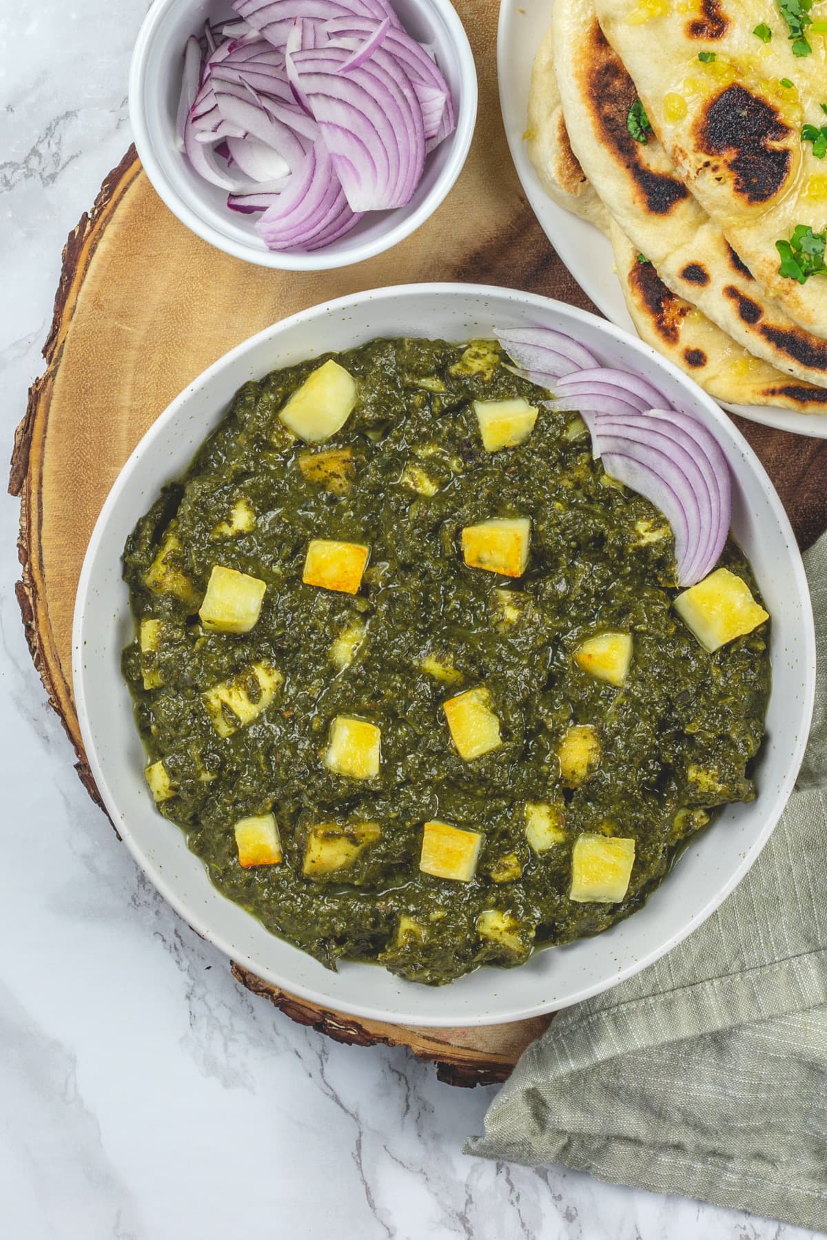 Saag paneer with sliced onions and naan on a wooden board.