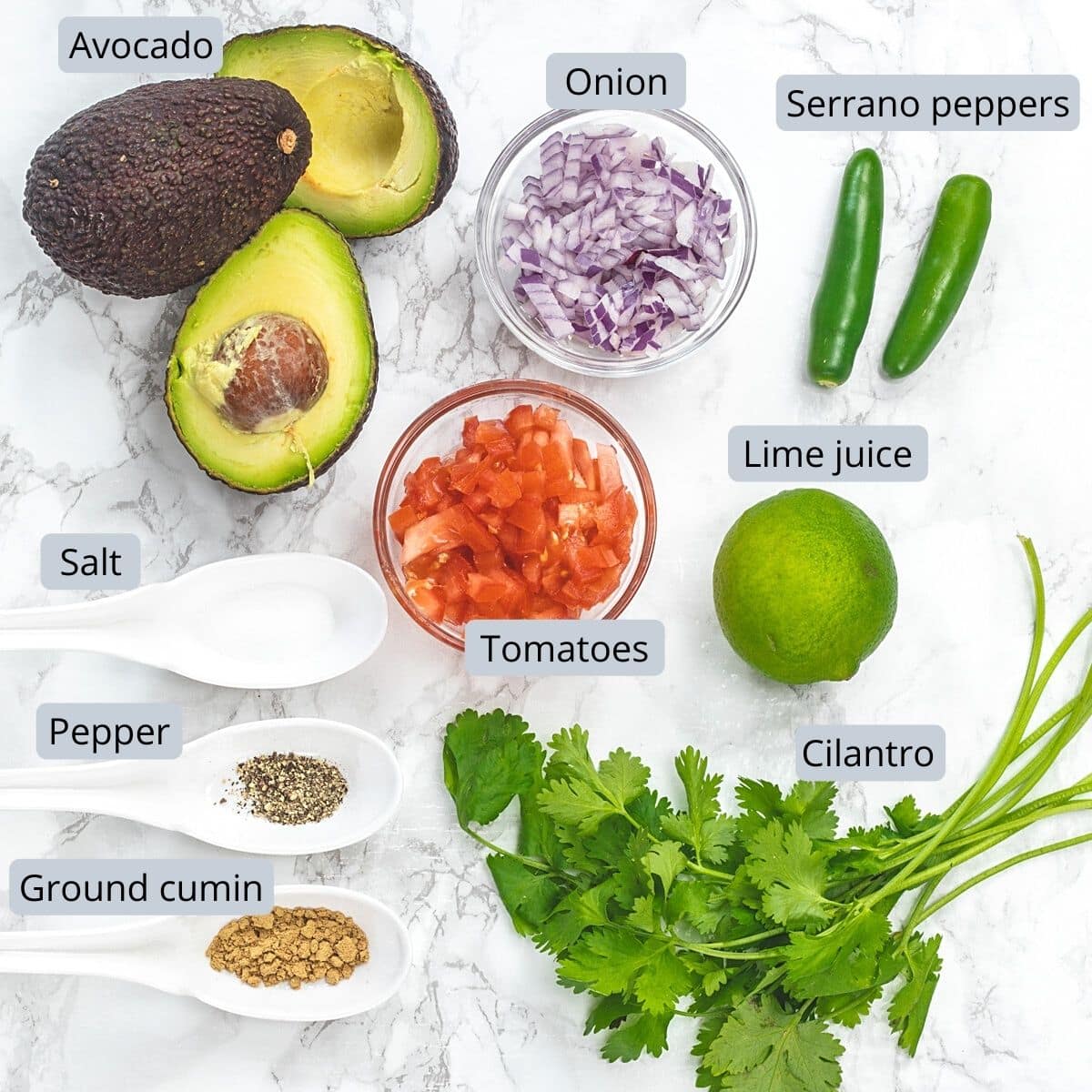 Ingredients for spicy guacamole in individual bowls, spoons and marble surface.