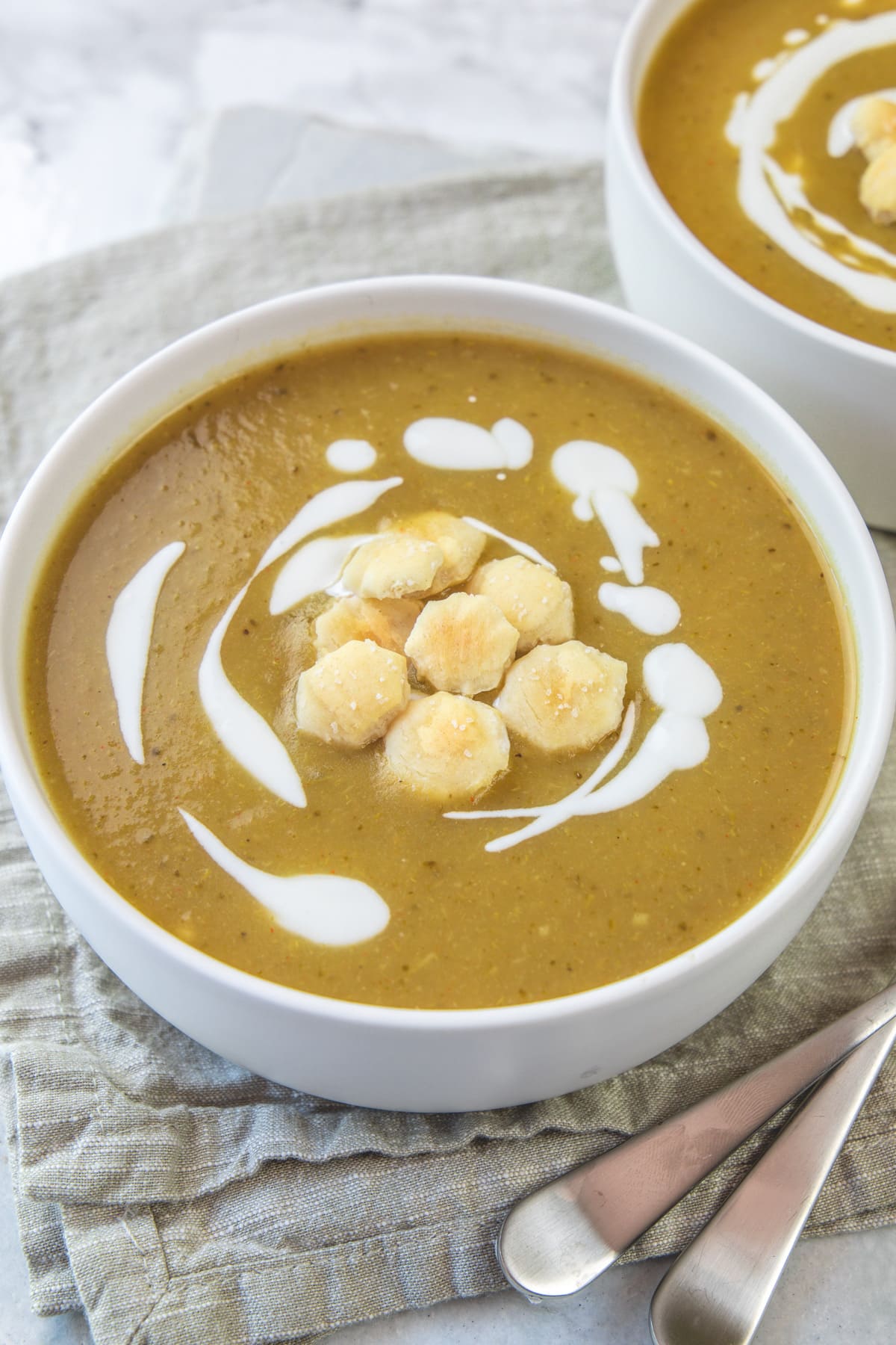 Close up of broccoli potato soup bowl with crackers and swirls of yogurt with spoons on side.