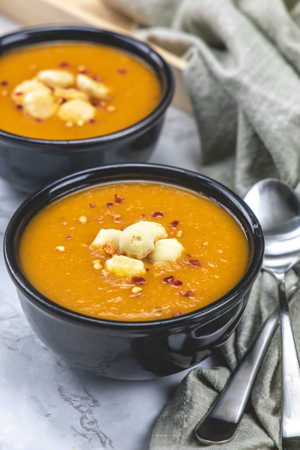 2 bowls of carrot tomato soup garnished with chili flakes, crackers with two spoons on side.