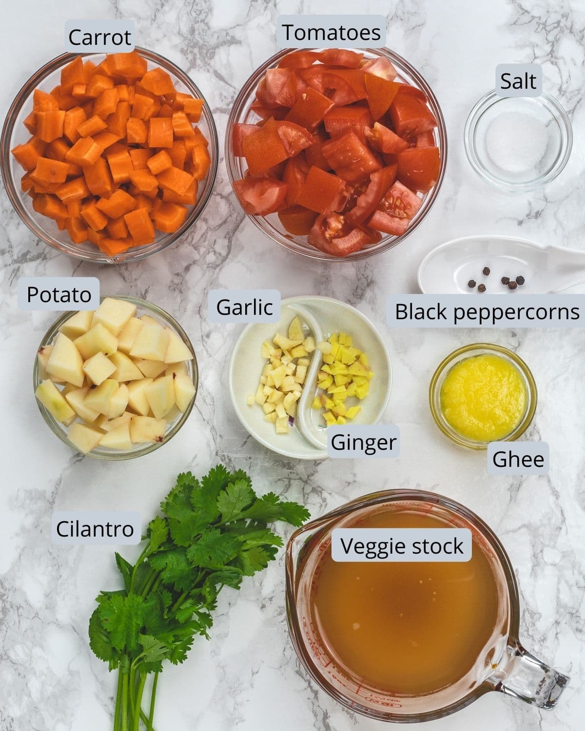 Carrot tomato soup ingredients in individual bowls on marble surface.