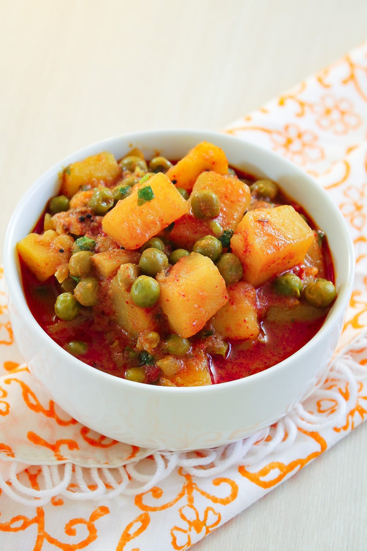 Aloo matar without onion garlic served in a white bowl with orange napkin under the bowl.