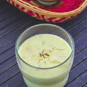 A glass of badam milk garnished with sliced almonds and saffron with another glass in the basked in back.