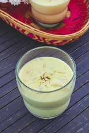 A glass of badam milk garnished with sliced almonds and saffron with another glass in the basked in back.