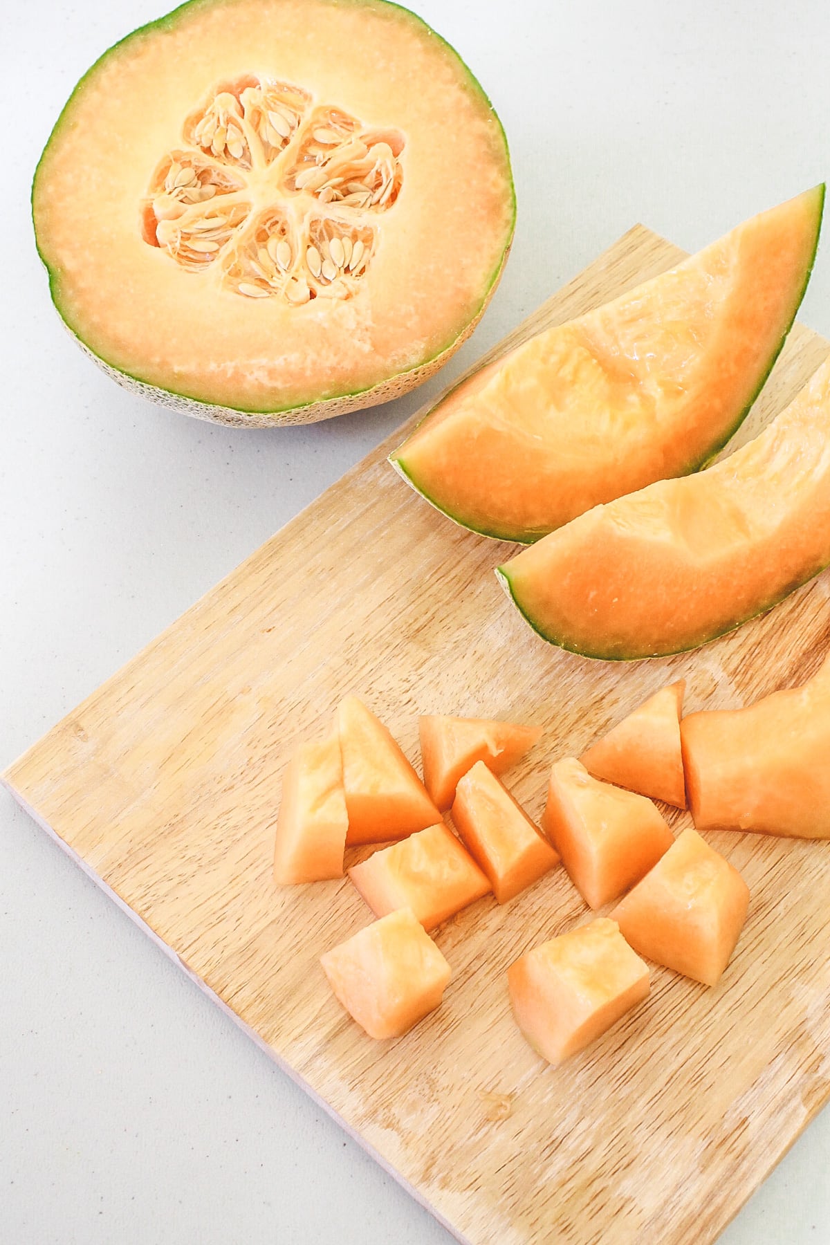 Halve cantaloupe, few slices and few cubed pieces on a wooden chopping board.