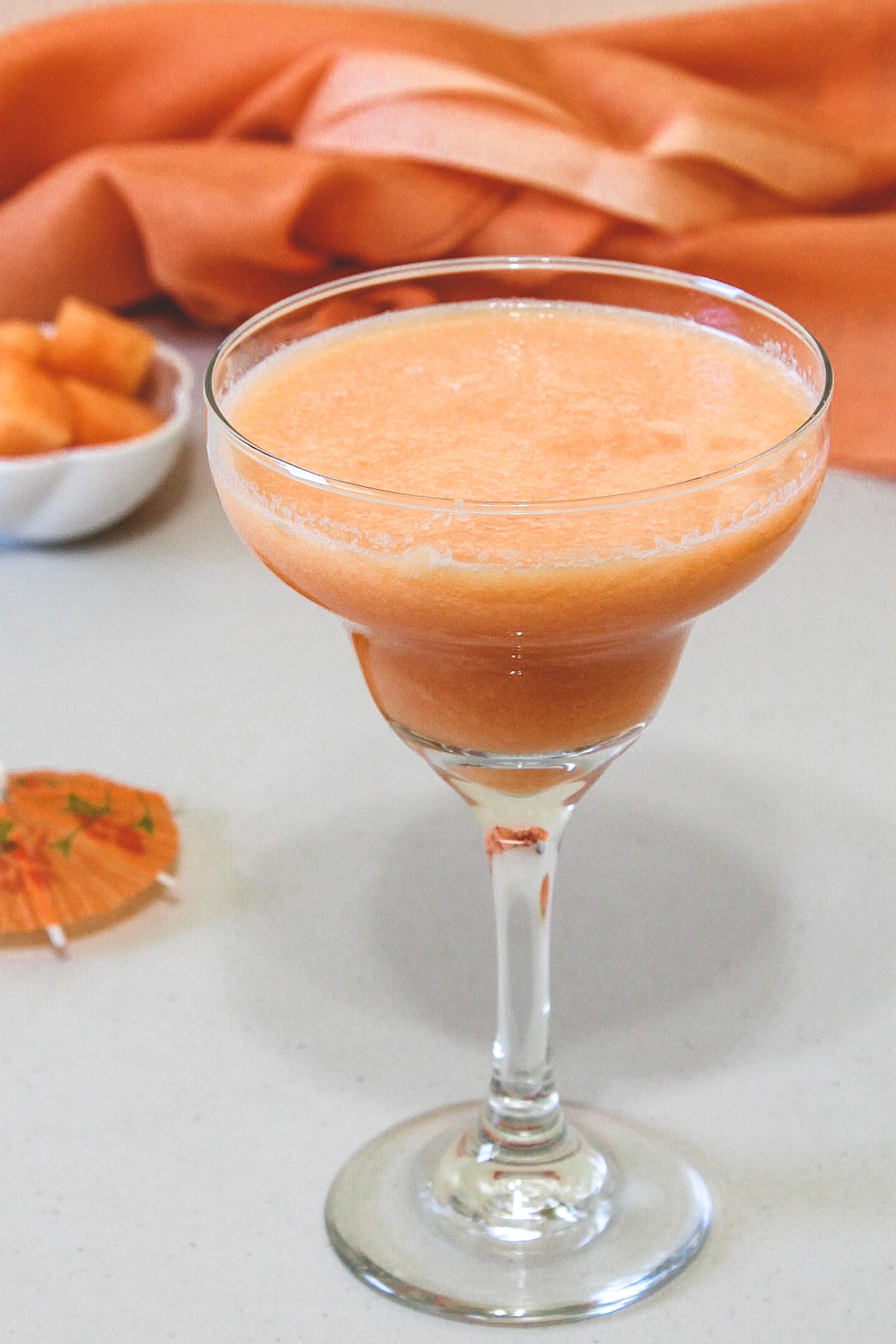 A glass of cantaloupe juice with cantaloupe cubes in a bowl in the back with orange fabric.