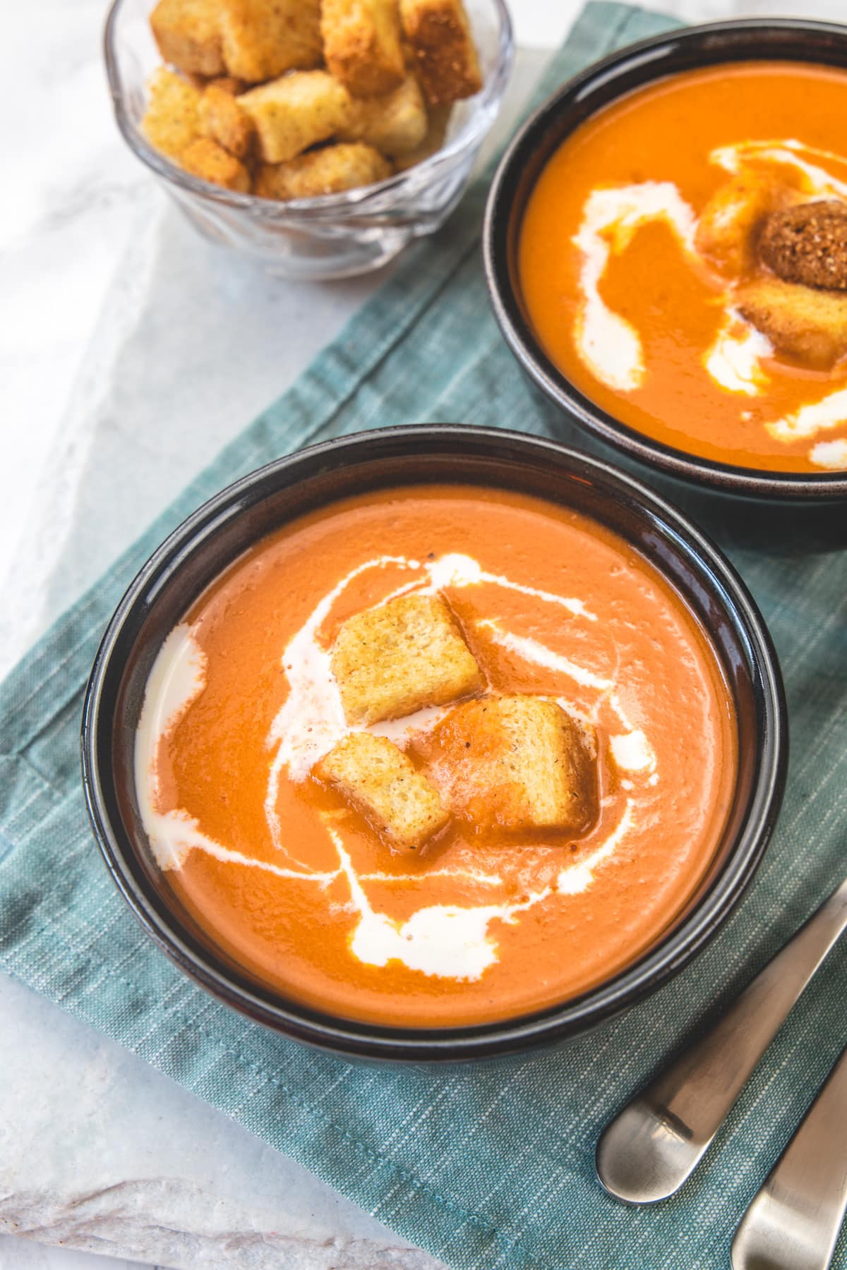 Bowls of tomato soup garnished with croutons and cream with spoons on side.