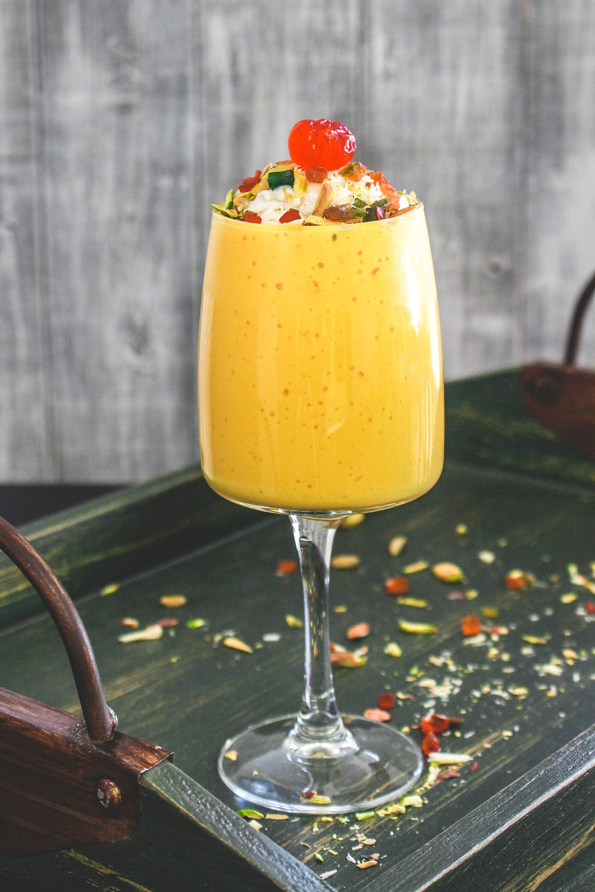 Glass of mango mastani in a tray with nuts spread around.
