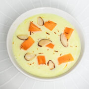Mango raita served in a bowl and plate underneath, raita garnished with almonds, mango and saffron.